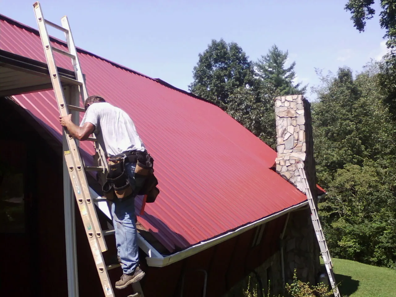 Roofer on a ladder beside a red metal roof and stone chimney — Soffit & Fascia Repair by Roofing Brothers in Little Plymouth