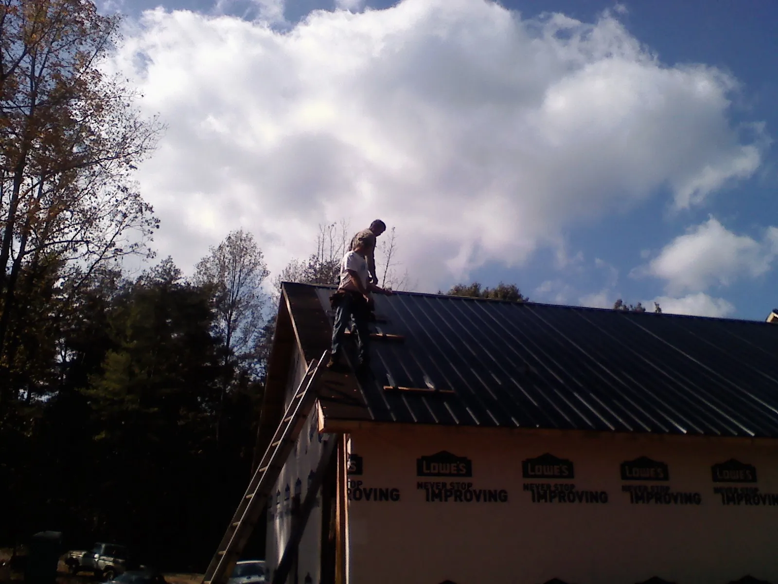 Roofer fastening dark metal panels high against a cloudy sky, from a Roof Inspection job in Little Plymouth